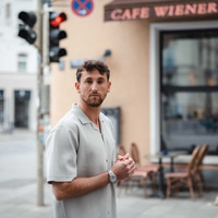 Mies kahvilan edessä, vaalea paita ja kello, katsoo kameraan.