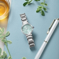 Mintunvihreä Citizen Eco-Drive -rannekello hopeanvärisellä rannekkeella lepää sinisellä kuvioidulla pinnalla valkoisen kynän ja meripihkanvärisen nesteen lasin vieressä.