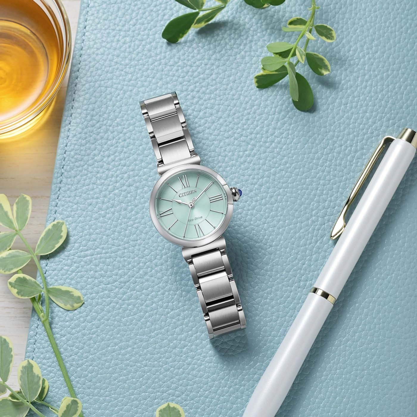 Mintunvihreä Citizen Eco-Drive -rannekello hopeanvärisellä rannekkeella lepää sinisellä kuvioidulla pinnalla valkoisen kynän ja meripihkanvärisen nesteen lasin vieressä.
