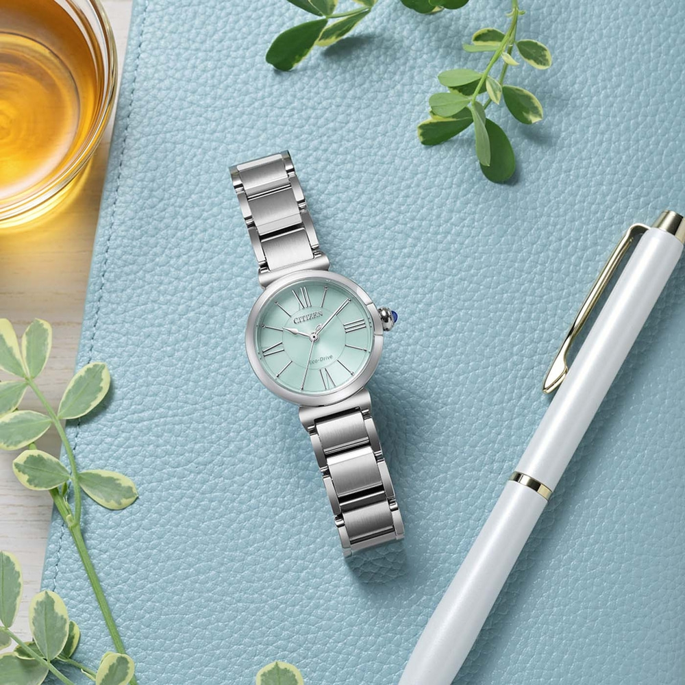Mintunvihreä Citizen Eco-Drive -rannekello hopeanvärisellä rannekkeella lepää sinisellä kuvioidulla pinnalla valkoisen kynän ja meripihkanvärisen nesteen lasin vieressä.