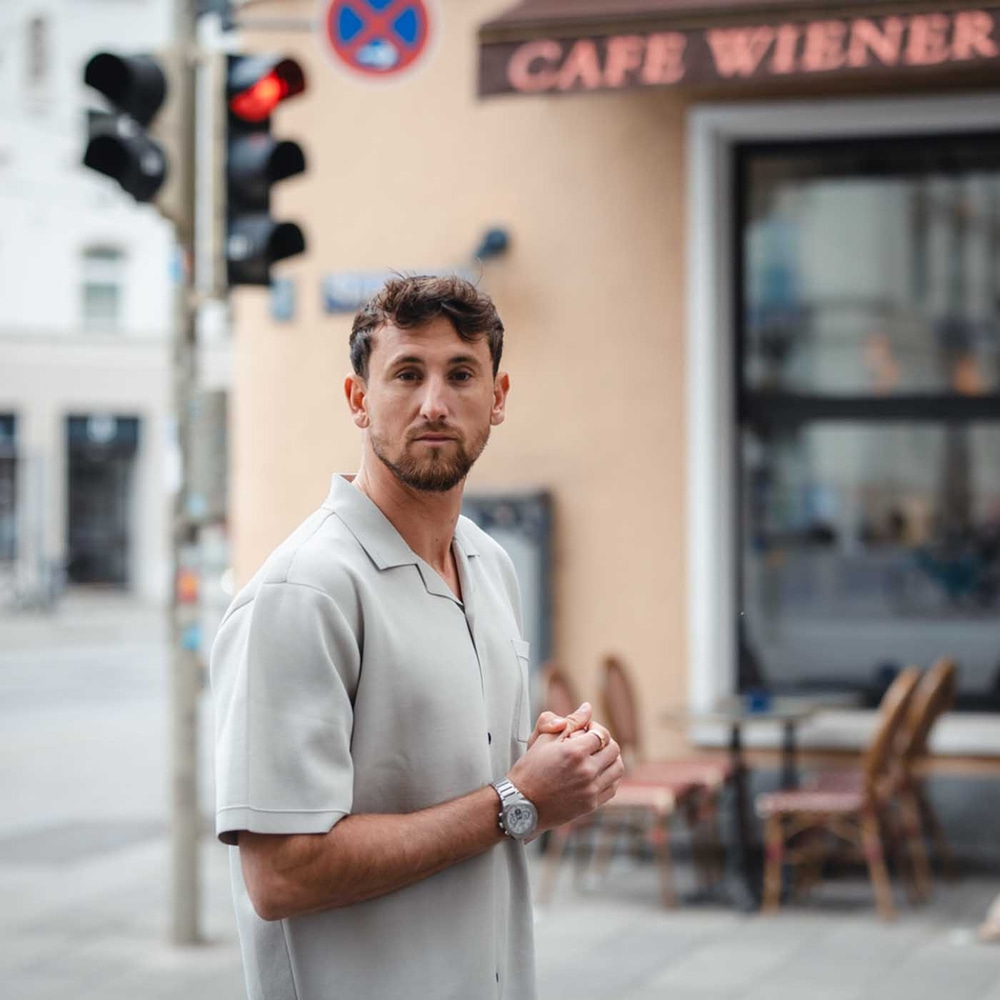 Mies kahvilan edessä, vaalea paita ja kello, katsoo kameraan.